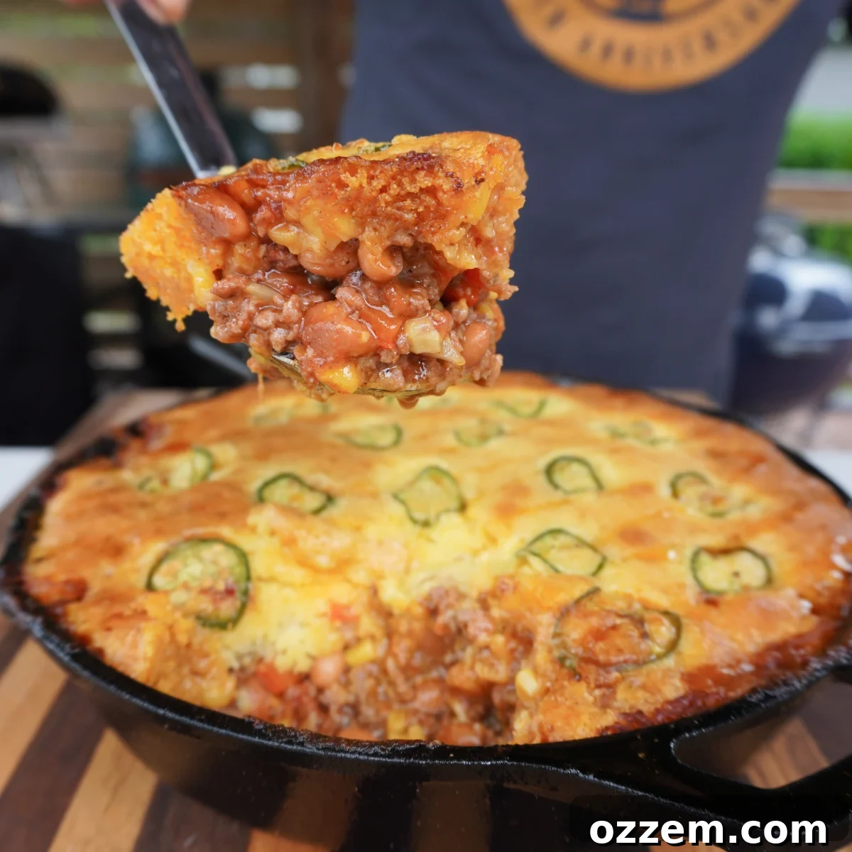 A perfectly smoked cornbread cowboy casserole, hot out of a cast iron skillet, with a spoonful removed to show its rich, layered interior of savory beef and vegetables beneath a golden cornbread crust.