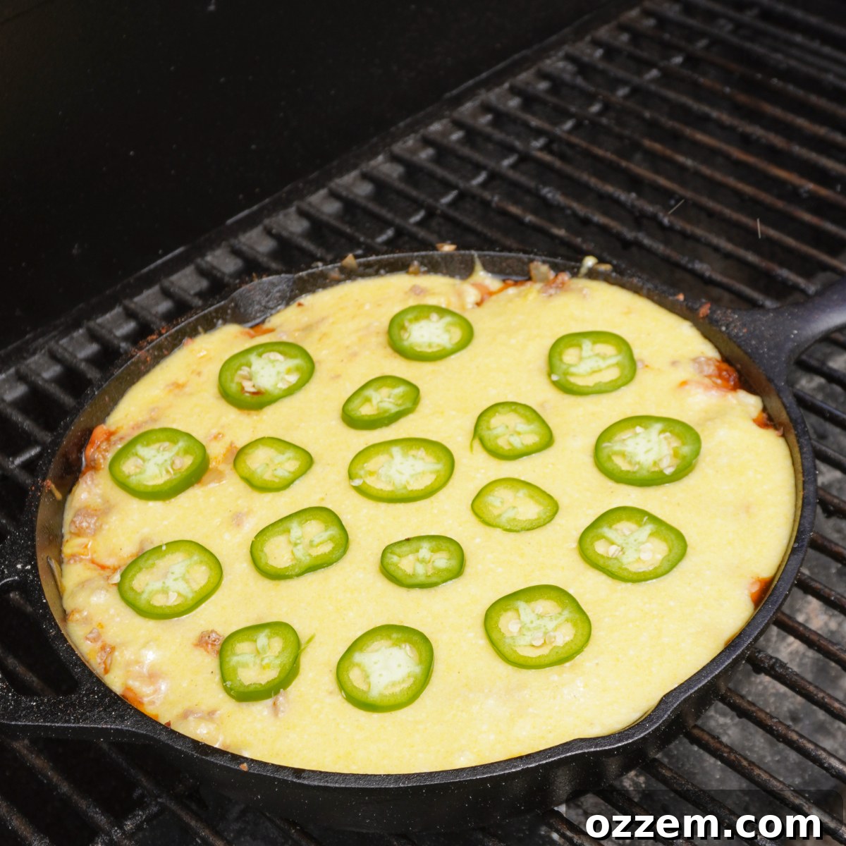 The assembled cornbread cowboy casserole, topped with a smooth layer of cornbread batter and garnished with sliced jalapenos, baking on the grill in a cast iron skillet.