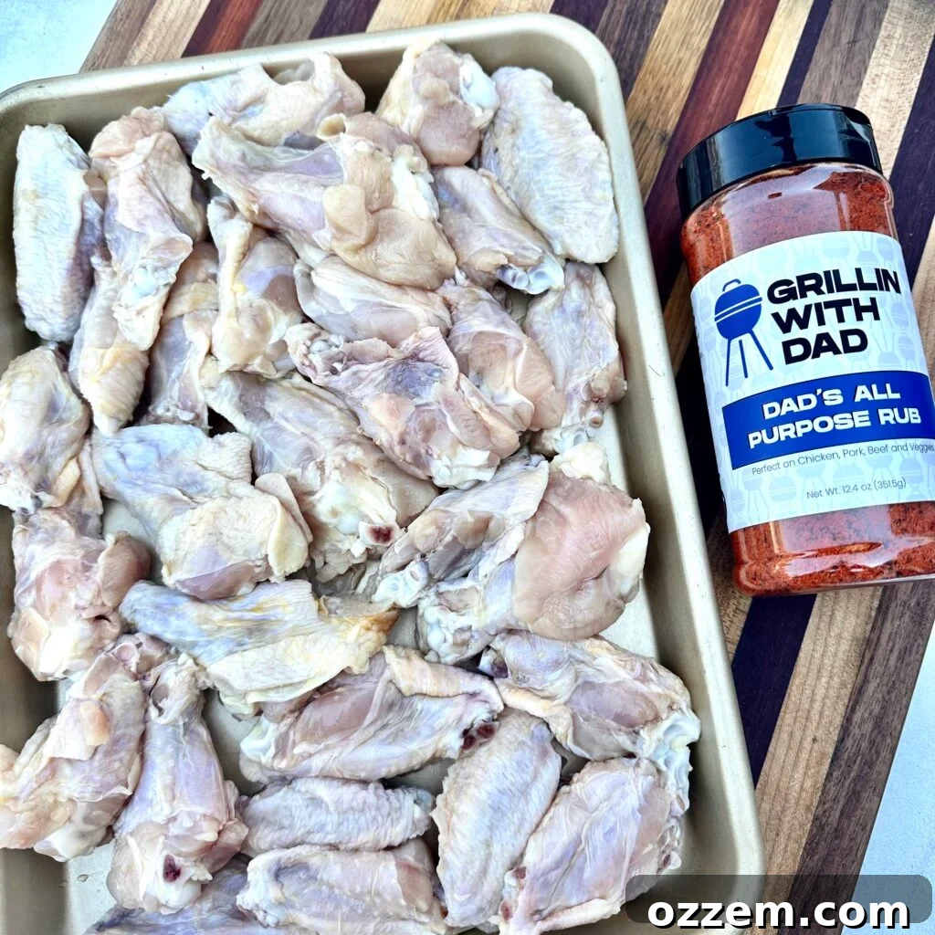 Raw party wings neatly arranged on a metal tray, with a bottle of Dad's All Purpose Rub positioned beside them, ready for a generous seasoning application.