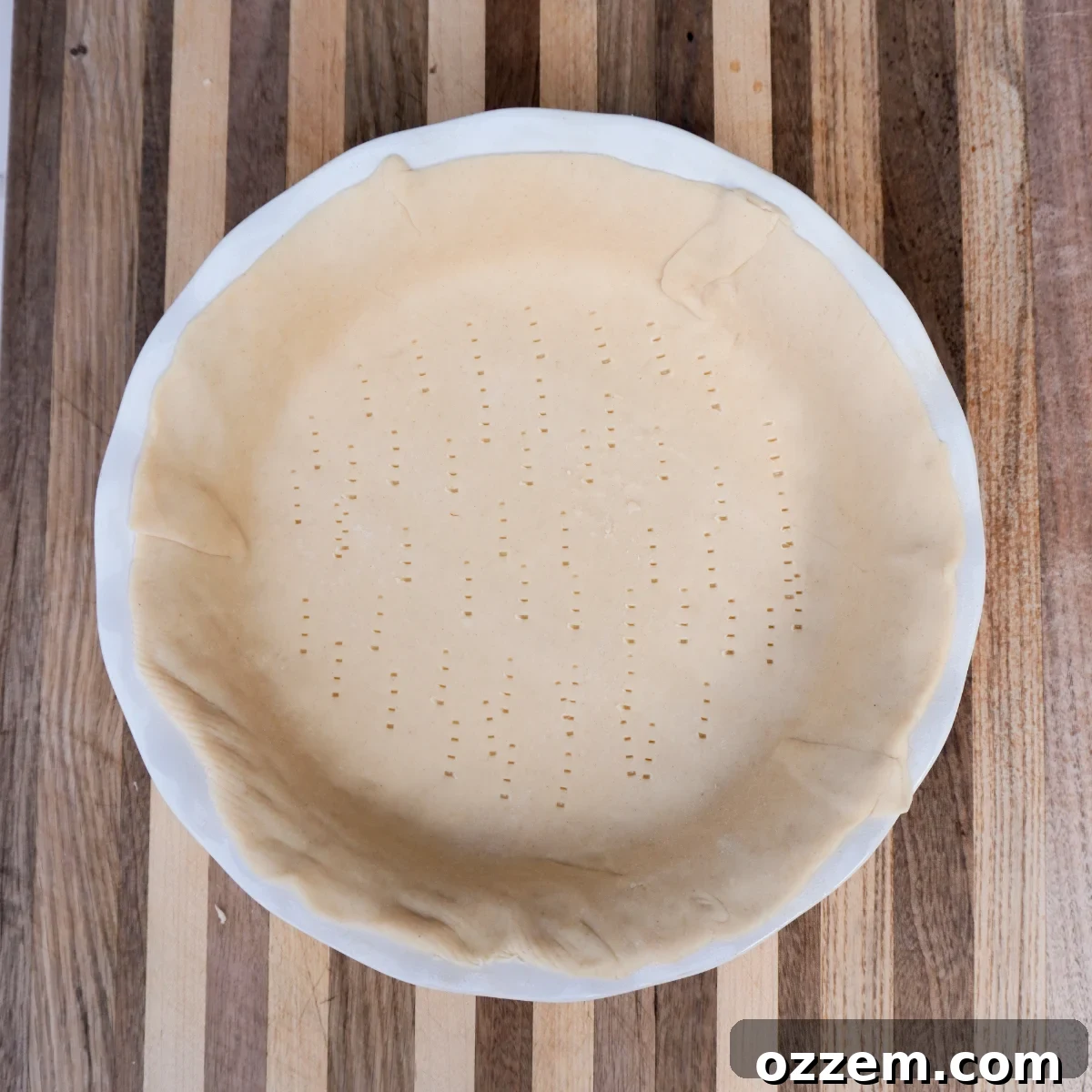 Raw pie crust neatly molded into a white ceramic pie dish, resting on a wooden cutting board, ready for blind baking.