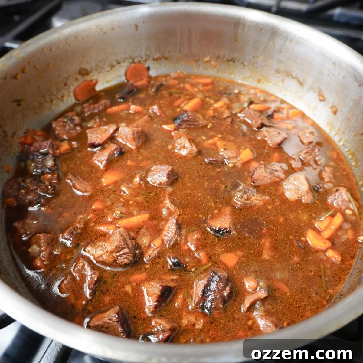 Rich, dark smoked meat pie gravy bubbling gently in a metal saucepan on the stove, thick with tender meat and vegetables.