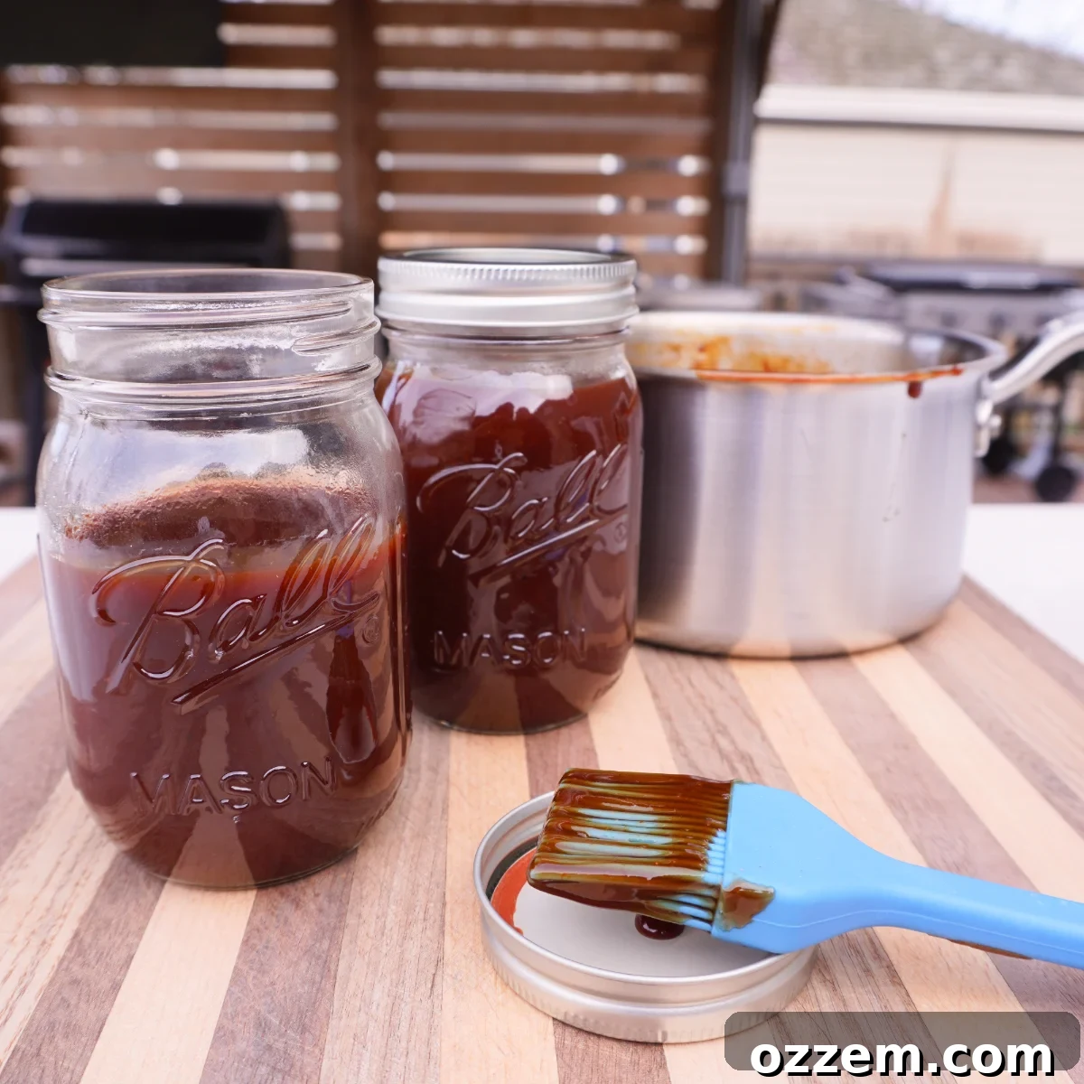 Multiple mason jars filled with homemade BBQ sauce arranged on a cutting board, alongside a saucepan and a dirty silicone brush.