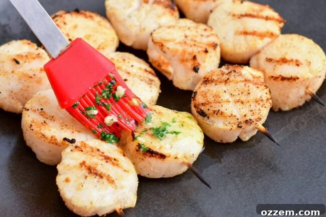 A hand using a brush to generously apply melted herb butter onto freshly grilled scallops on a plate.