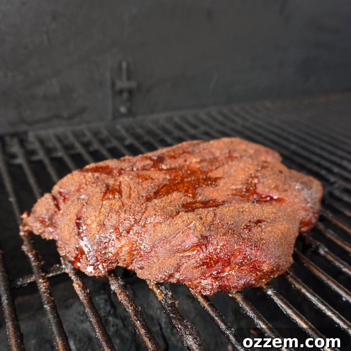 A seasoned chuck roast resting on the grates of a smoker, acquiring a rich, dark bark.