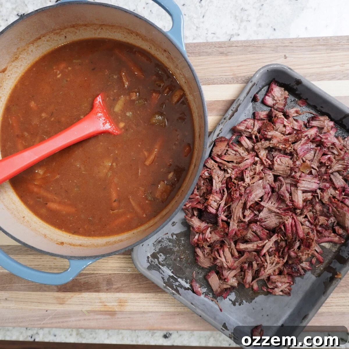 Shredded beef placed next to the rich braising liquid and cooked vegetables in a Dutch oven.