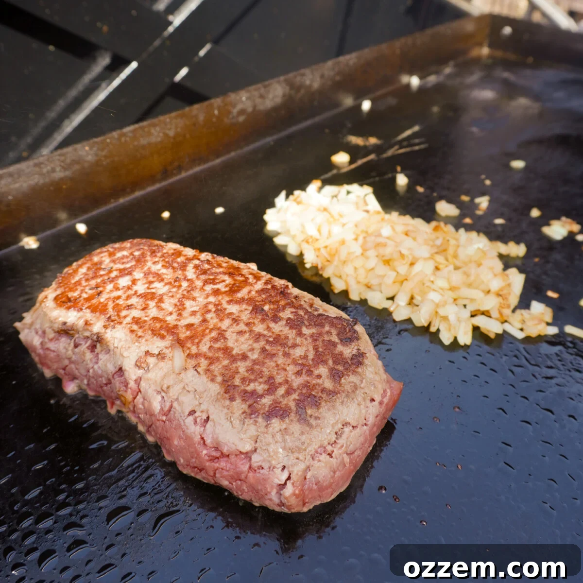 A block of ground beef and diced onions sizzling on a hot griddle, beginning to brown and caramelize.