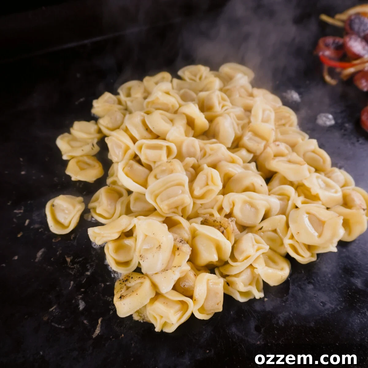 Frozen pre-cooked tortellini steaming under a basting dome on the griddle, ready to be mixed with grilled sausage and vegetables.