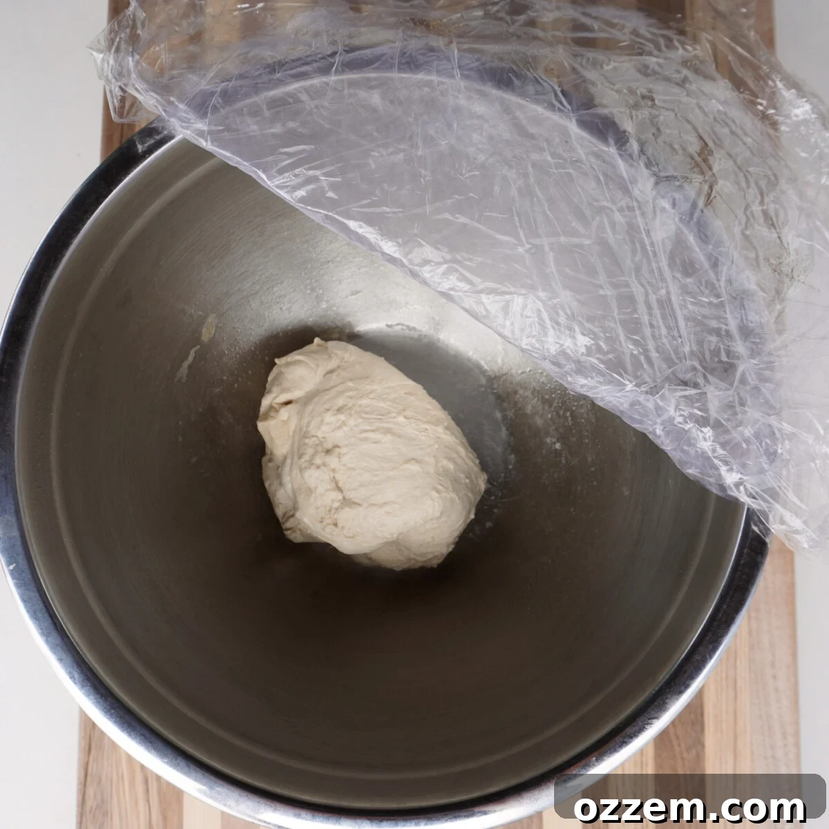 Homemade pizza dough undergoing its first rise, covered with plastic wrap, showing significant expansion.