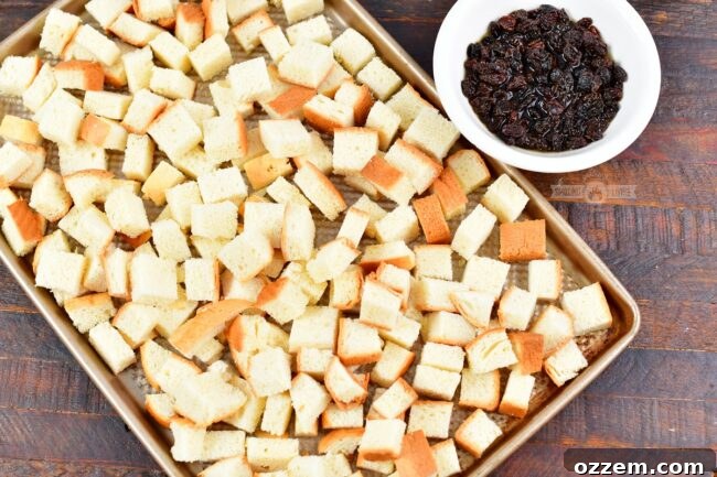 Uniformly cubed brioche bread spread on a baking sheet to stale, alongside a small bowl of dark bourbon-soaked raisins.