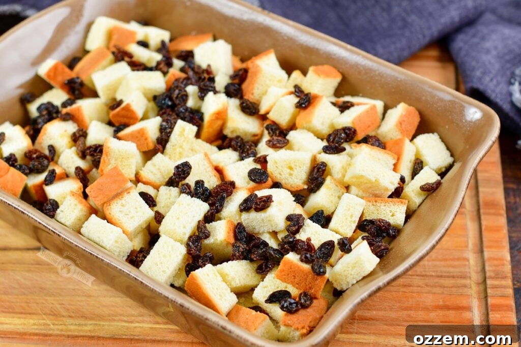 Cubed brioche bread and bourbon-soaked raisins spread evenly in a 9x13 inch baking dish, ready for the custard mixture.
