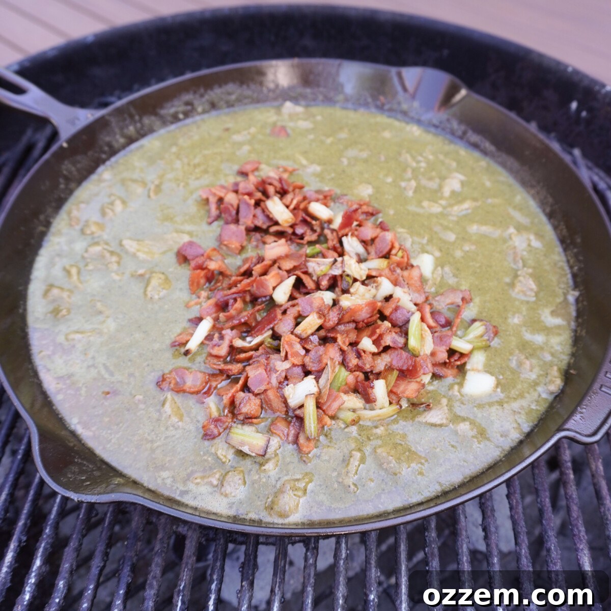 A cast iron skillet filled with simmering Grilled Carne en su Jugo, rich with beef, bacon, and salsa verde, cooking over indirect heat on a charcoal grill.