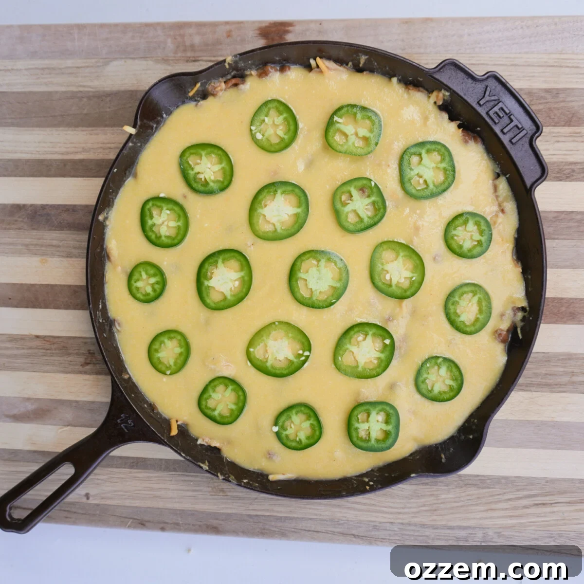 A pristine image depicting the smooth, meticulously prepared cornbread batter being poured with care and precision evenly over the chicken and bean mixture already settled in a rustic cast iron skillet, now perfectly poised for baking.