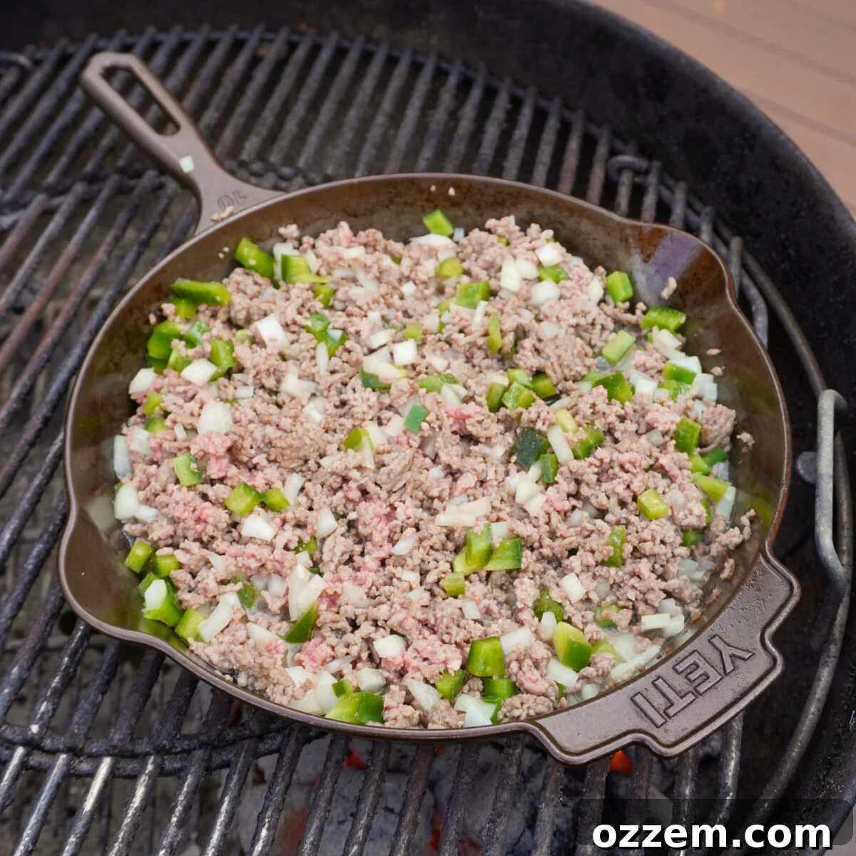 A cast iron skillet on the grill with browning ground beef and chopped vegetables.