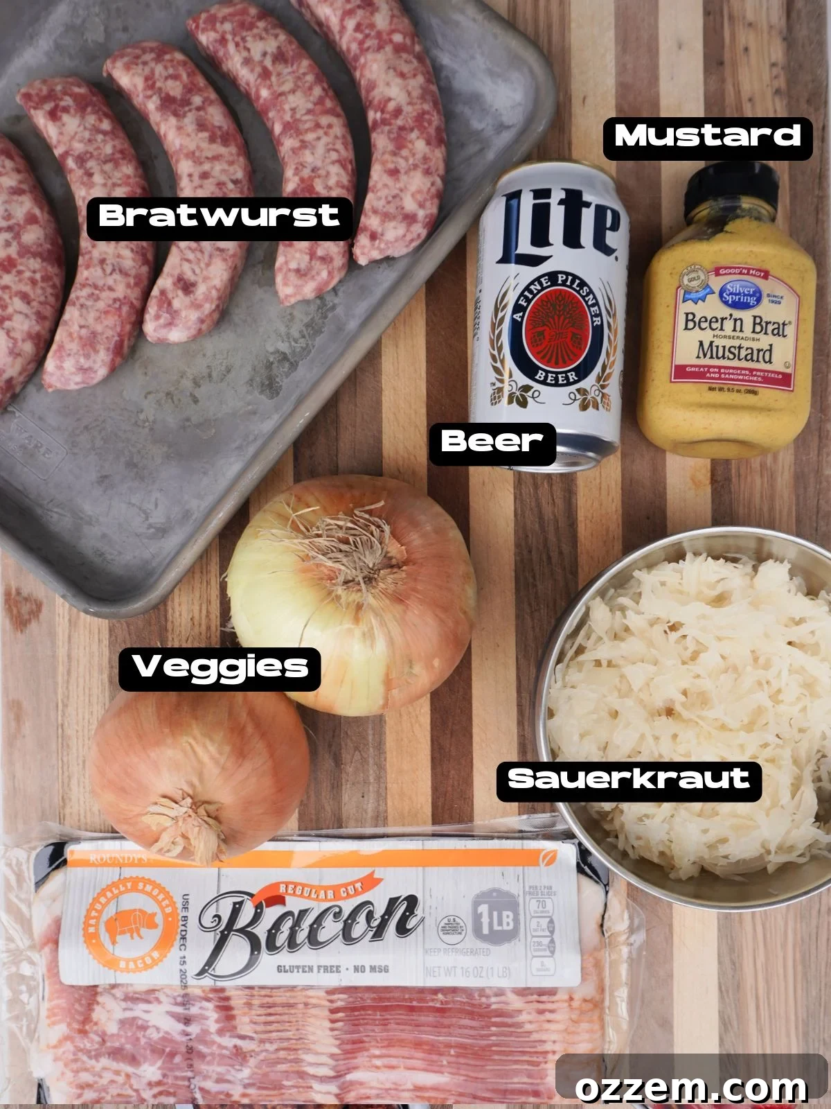 A variety of ingredients including bratwurst, onions, beer, sauerkraut, and mustard laid out on a wooden board.