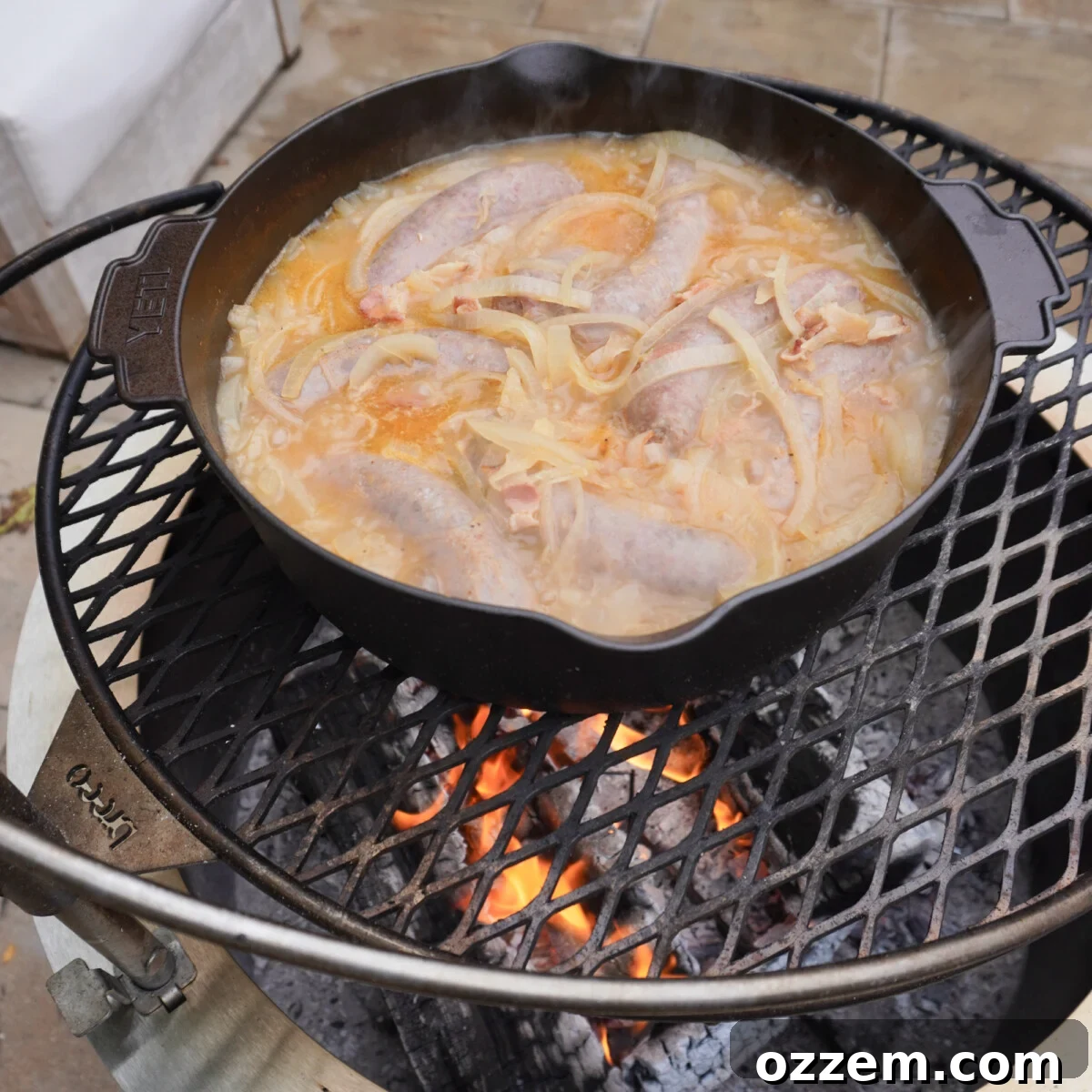 A Dutch oven containing beer brats and onions simmering on a Breeo wood-burning grill over an open fire.