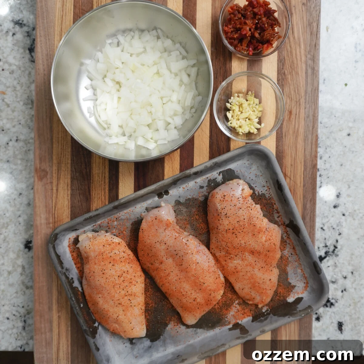 Raw chicken breasts seasoned with all-purpose rub, next to chopped onions and sun-dried tomatoes.