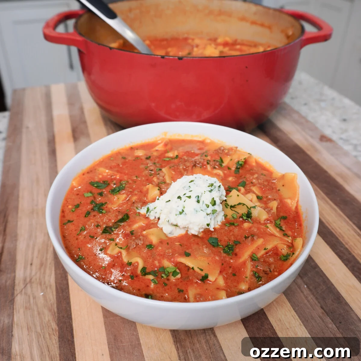 Lasagna soup plated in a white bowl on a stripped cutting board with ricotta cheese on top.
