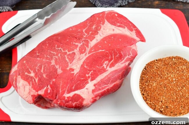 A raw beef chuck roast generously coated with seasoning rub, resting on a cutting board, with a bowl of beef rub nearby