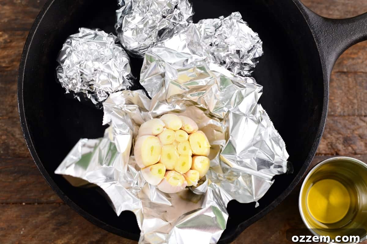 Multiple garlic bulbs wrapped in foil, drizzled with oil, and placed in a skillet.
