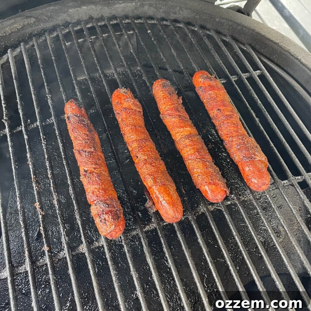 Raw beef hot dogs expertly wrapped in bacon and generously seasoned with BBQ rub, ready for grilling.