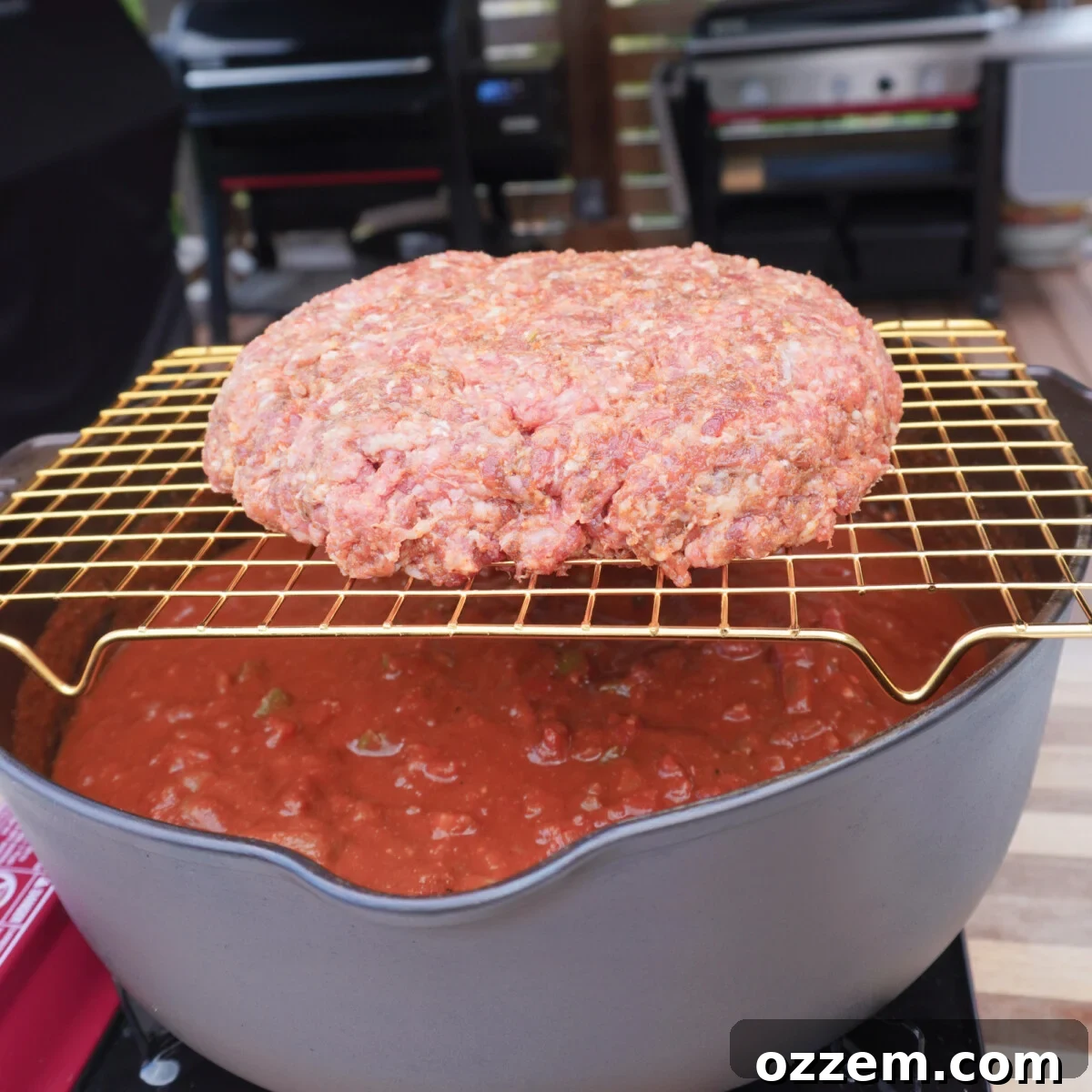 A seasoned beef patty resting on a wire rack, positioned directly over a Dutch oven filled with chili mix, ready for smoking.