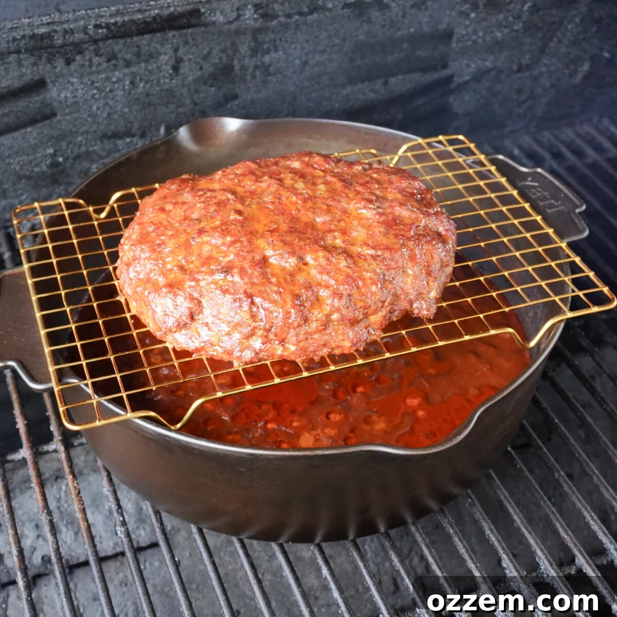 A Dutch oven filled with chili base, with a seasoned beef patty on a wire rack suspended above it, smoking on a grill.