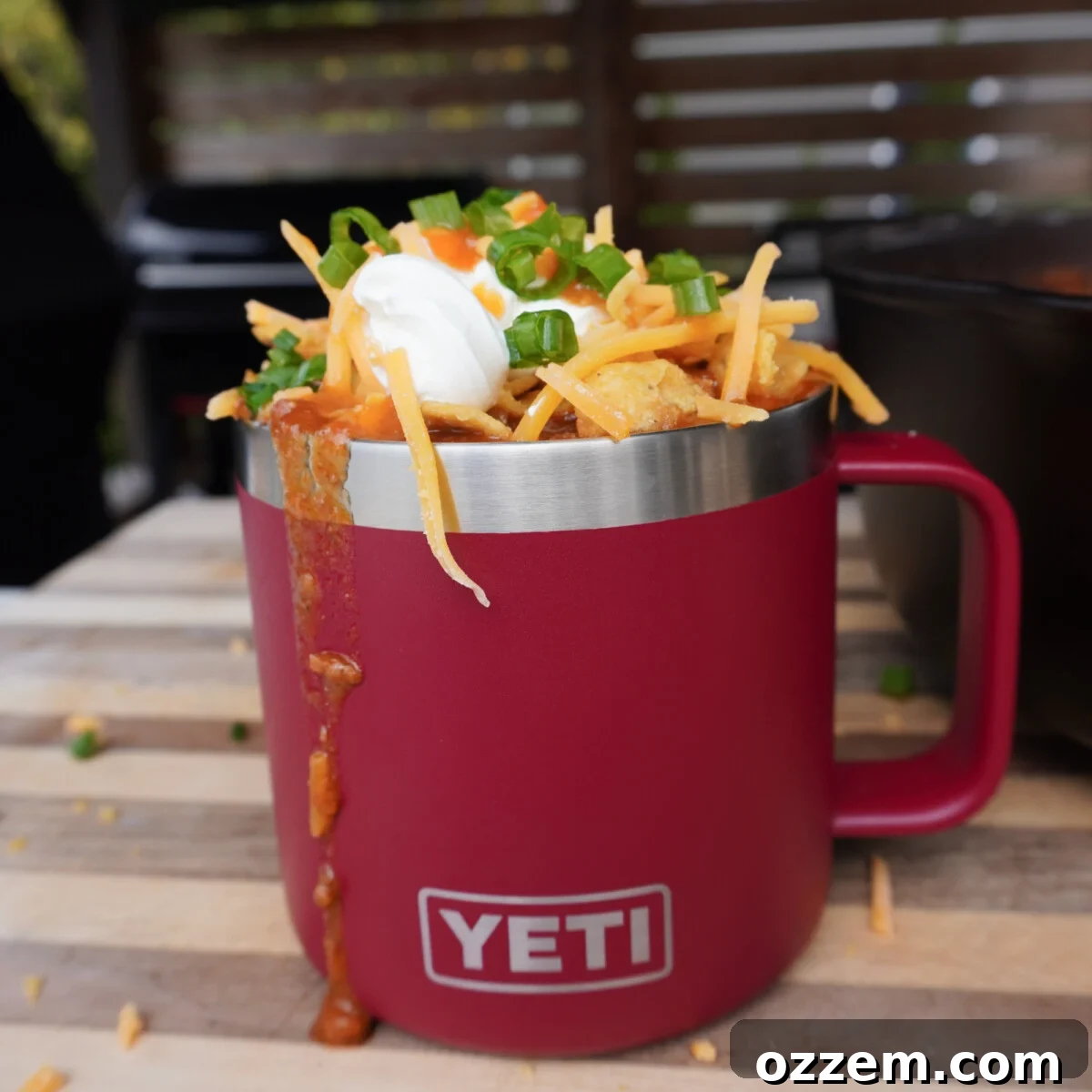 A red mug overflowing with Over the Top Beef Chili, garnished with shredded cheese, Fritos, and a dollop of sour cream.