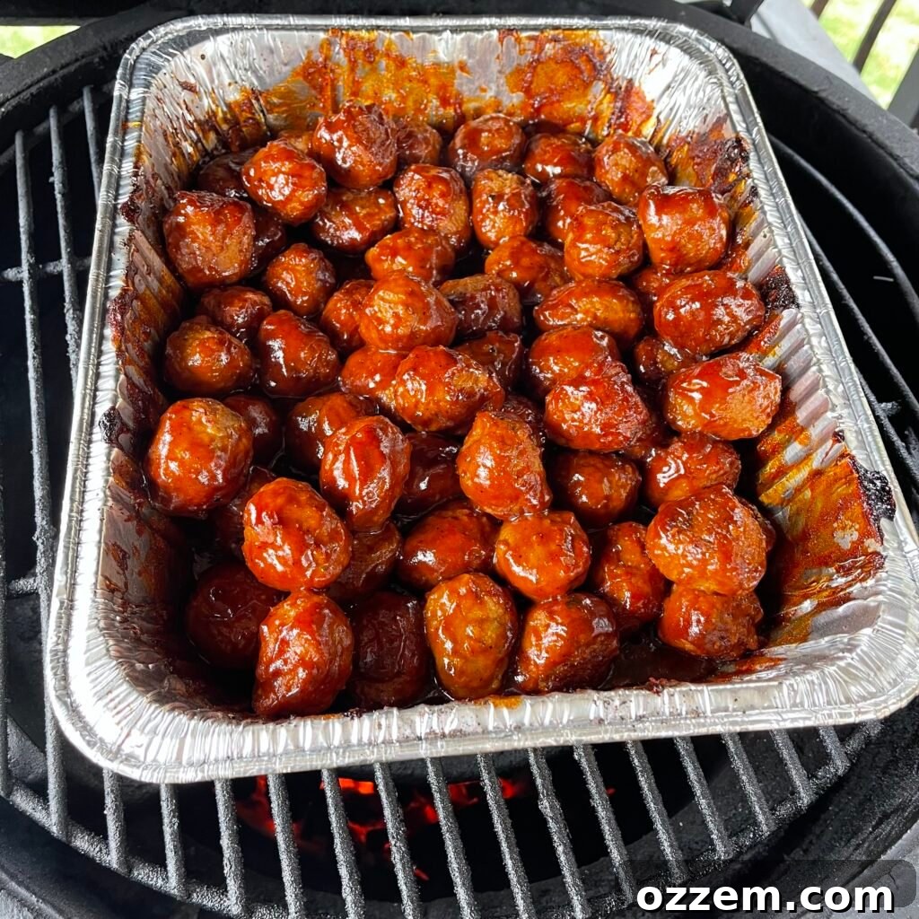 PB&J BBQ Meatballs slow cooking in an aluminum pan on a grill.