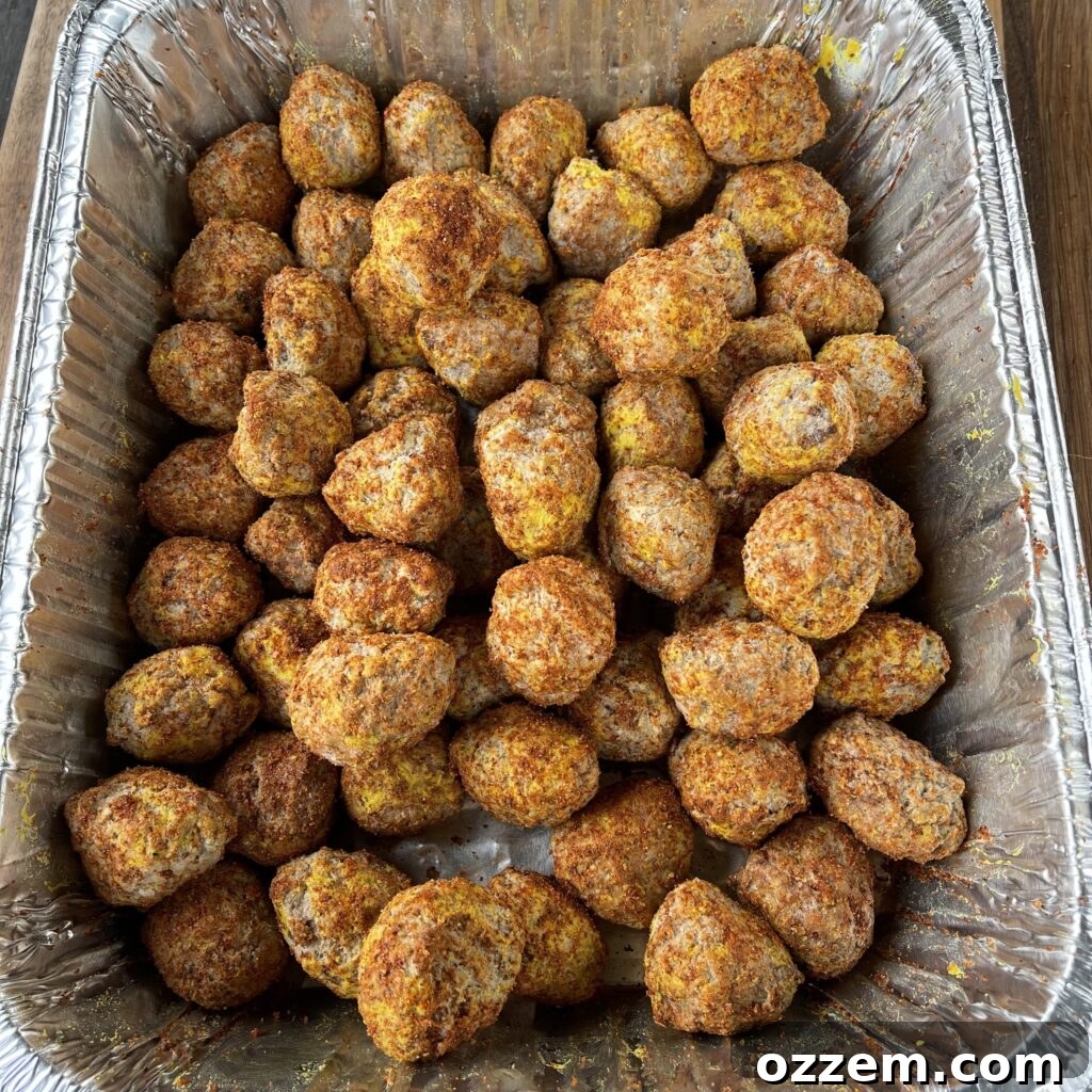 Frozen meatballs being coated with a yellow mustard binder and a savory BBQ rub in a disposable foil pan.
