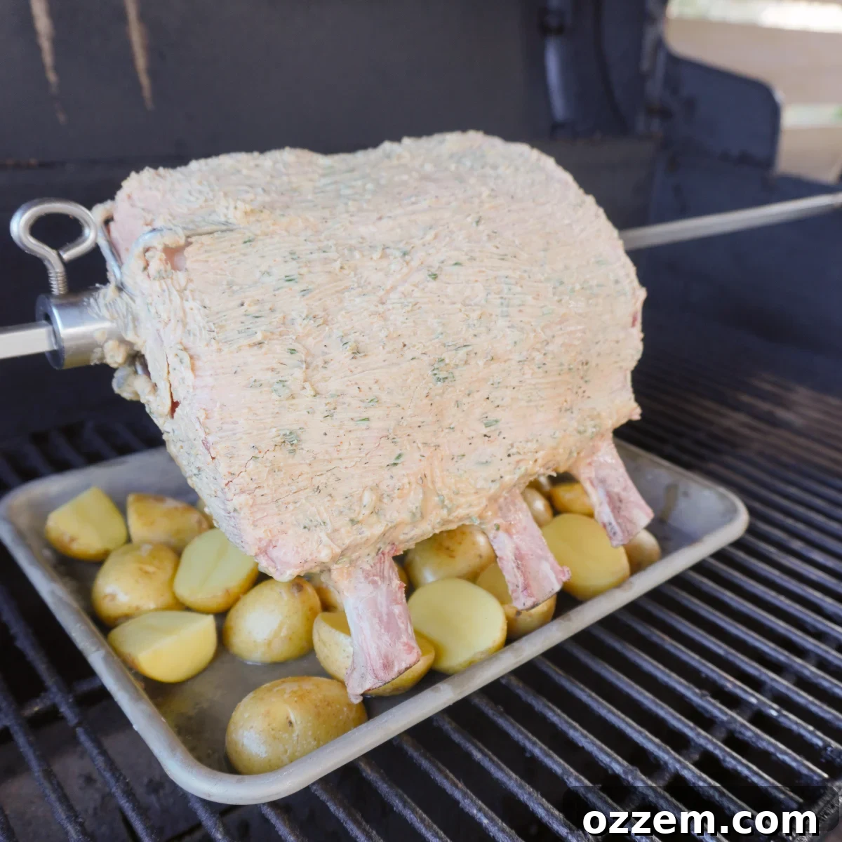 Seasoned and butter-spread raw prime rib skewered on the rotisserie, positioned above sliced potatoes on the grill, ready for cooking.