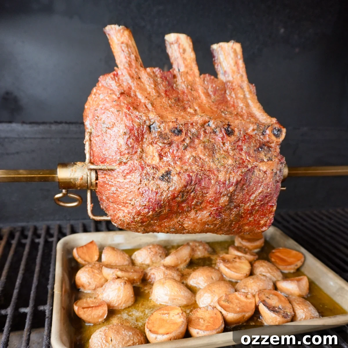 Cooked prime rib still on the rotisserie above sliced potatoes on the grill, showing its golden-brown crust.