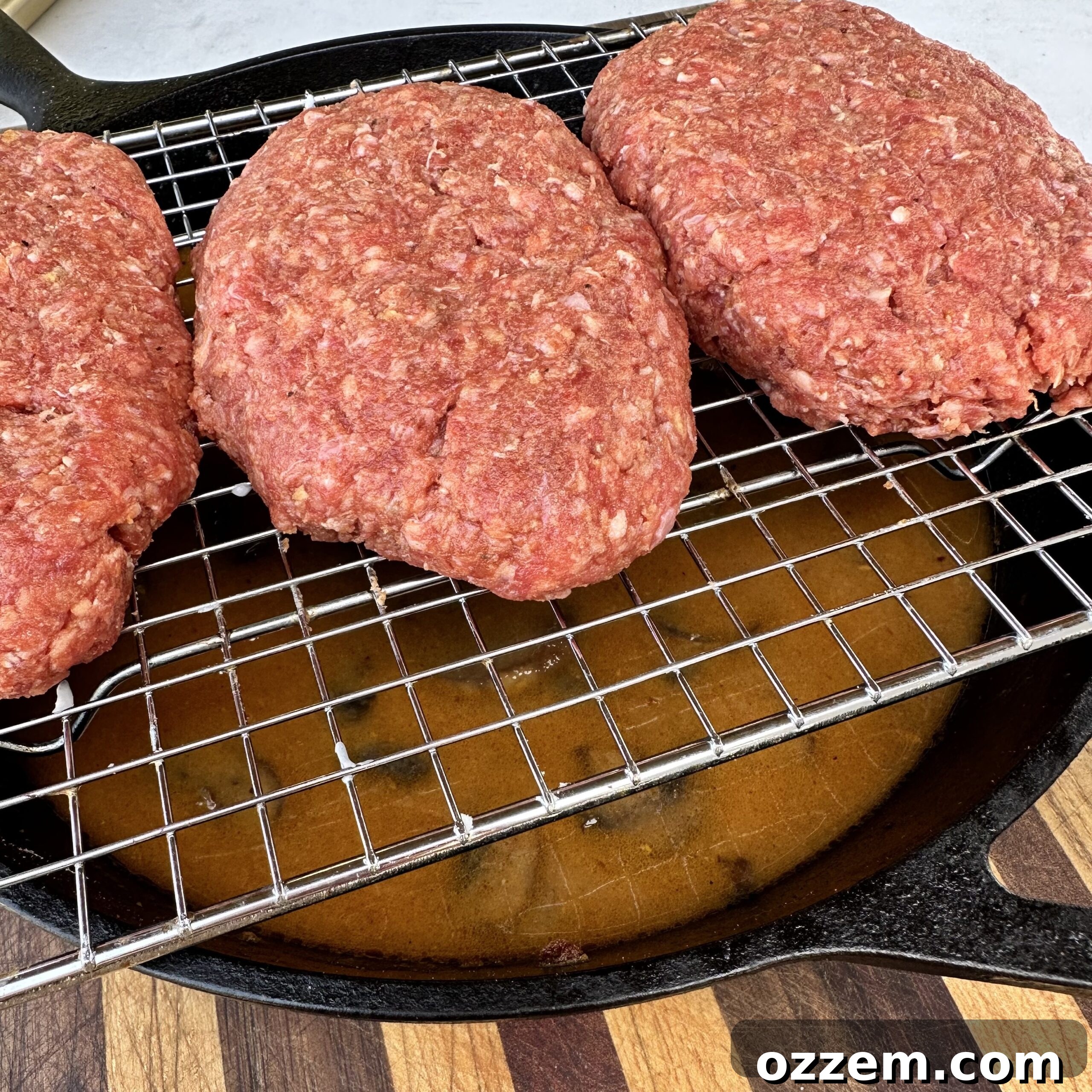 Salisbury steak patties cooking on a wire rack over a cast iron skillet with gravy on a grill.