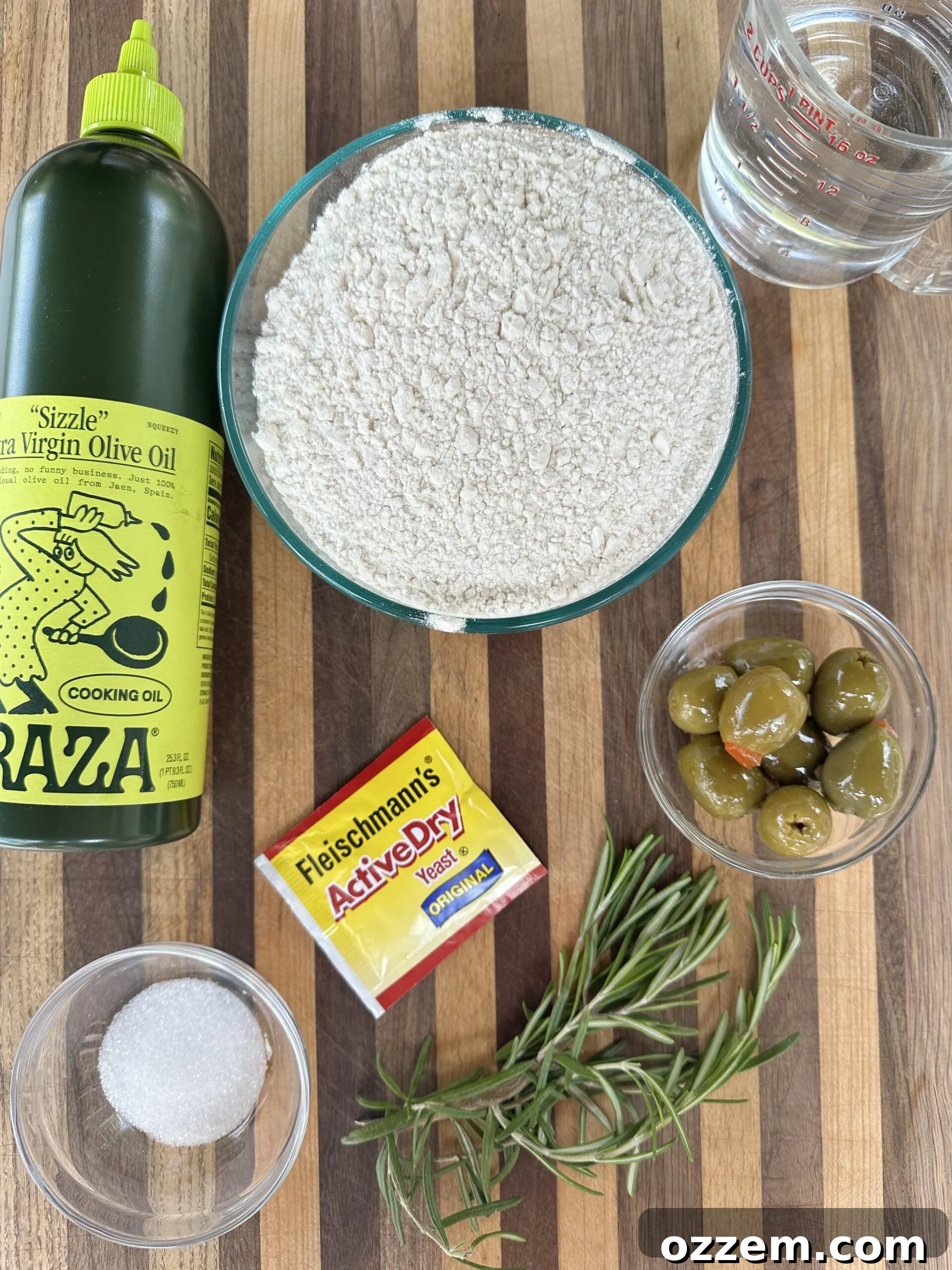 A collection of Focaccia bread ingredients: bread flour, olive oil, yeast, salt, honey, and a glass of water on a wooden surface.