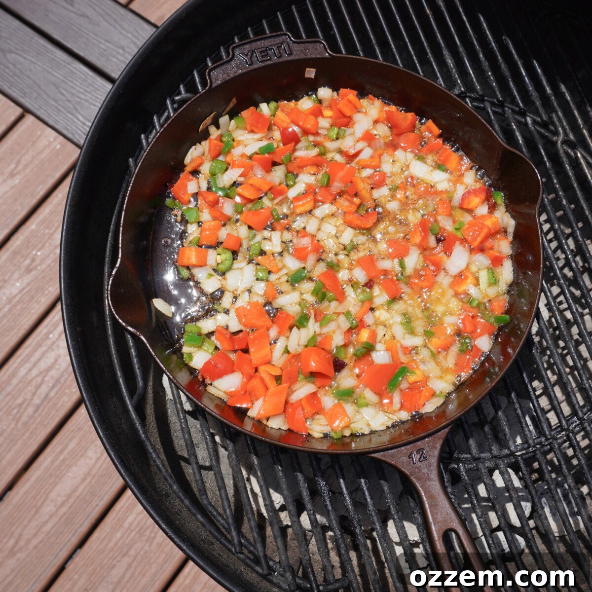 Sautéed onions, bell peppers, and jalapenos cooking in bacon grease within a cast iron skillet on the grill.