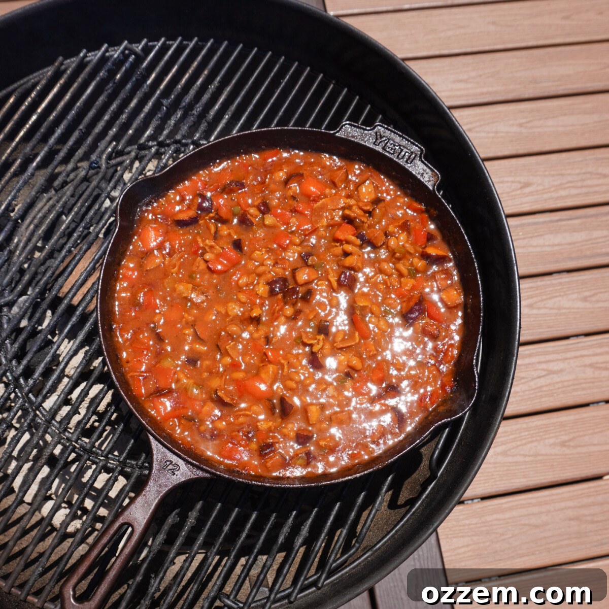 A cast iron skillet filled with mixed smoked pit beans, simmering on a charcoal grill with smoky embers.