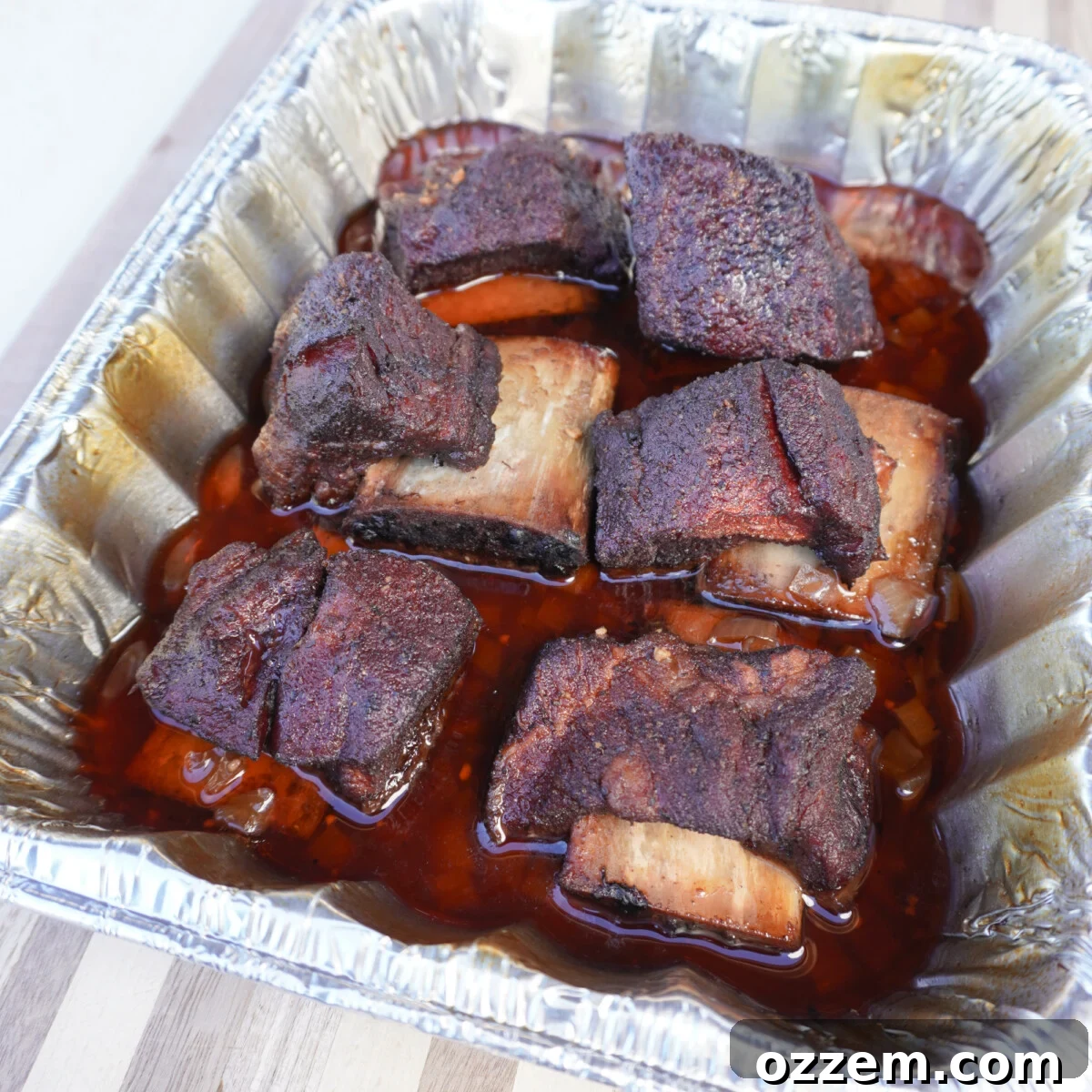 Velvet Smoke Ribs 12 Succulent cooked short ribs resting in a foil pan, glistening with rich braising liquid, ready for plating.