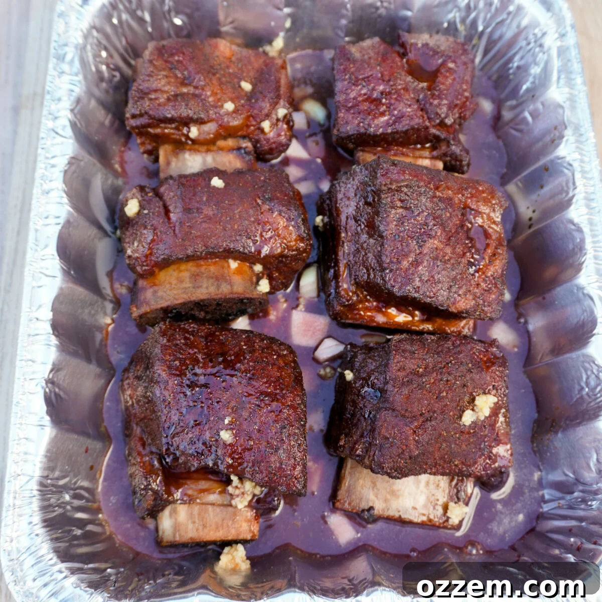 Velvet Smoke Ribs 10 Beef short ribs in a foil pan, generously covered with diced onions, beef stock, and red wine, ready for braising.