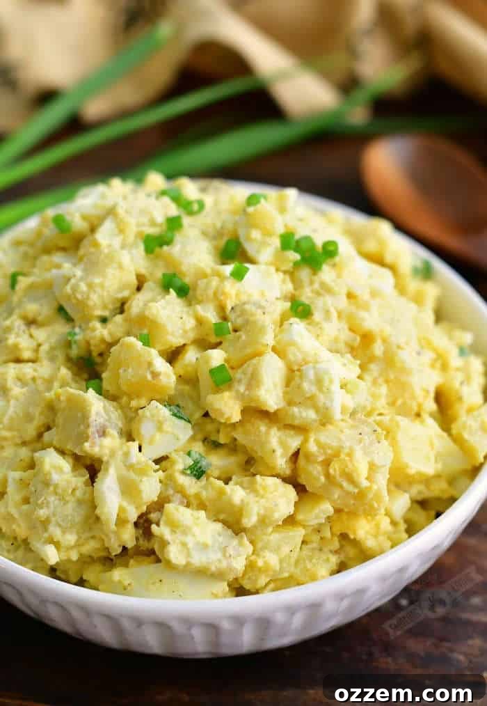 mixed potato salad in a white bowl topped with diced chives and a pooden spoon and green onion shoots on the background