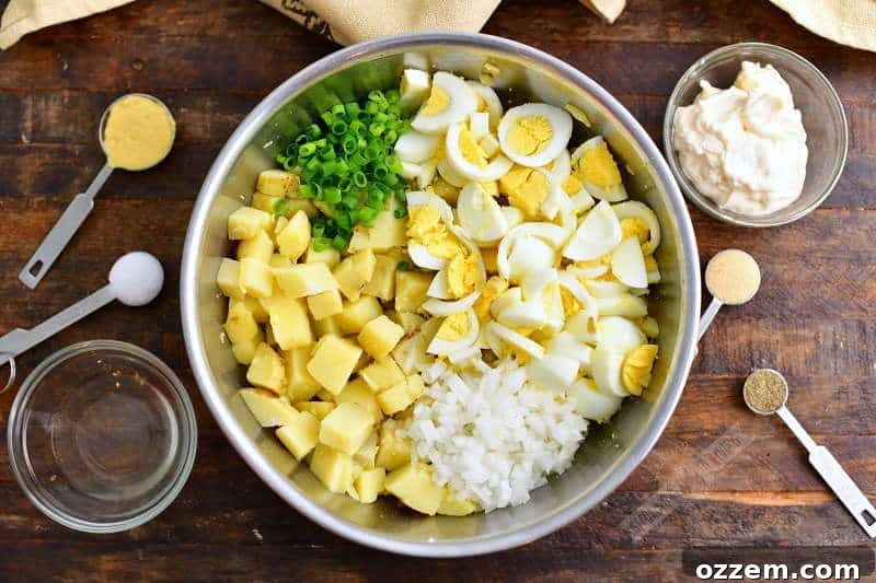 chopped potatoes, eggs, onion, and green onion in a metal bowl with a small bowl of mayo and vinegar next to it and measuring spoons of mustard, salt, pepper, and garlic powder next to the mixing bowl