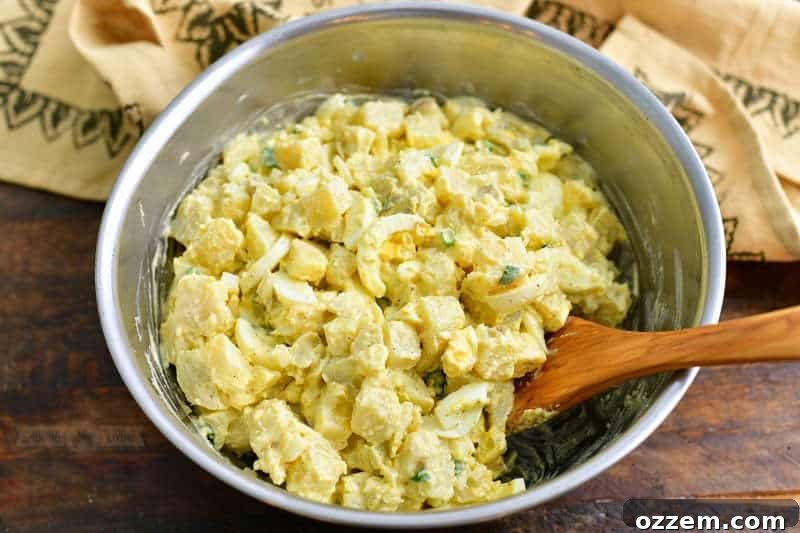 mixing potato salad ingredients together in a metal mixing bowl with a light brown towel behind it