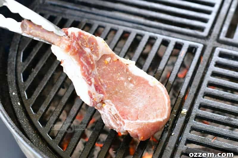 A raw, marinated pork chop being carefully placed onto a hot grill grate using tongs.