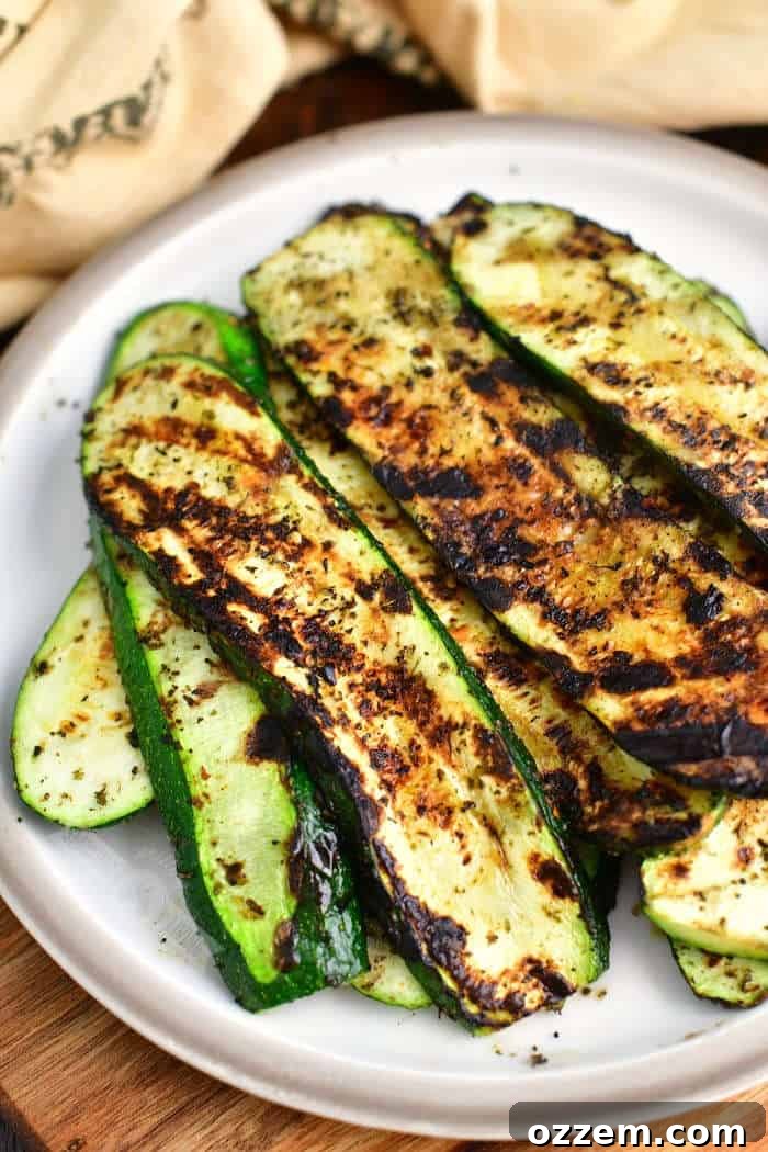 Long slices of perfectly grilled zucchini, golden-brown with distinct grill marks, arranged neatly on a white plate, with a light brown linen towel artfully placed beside it.