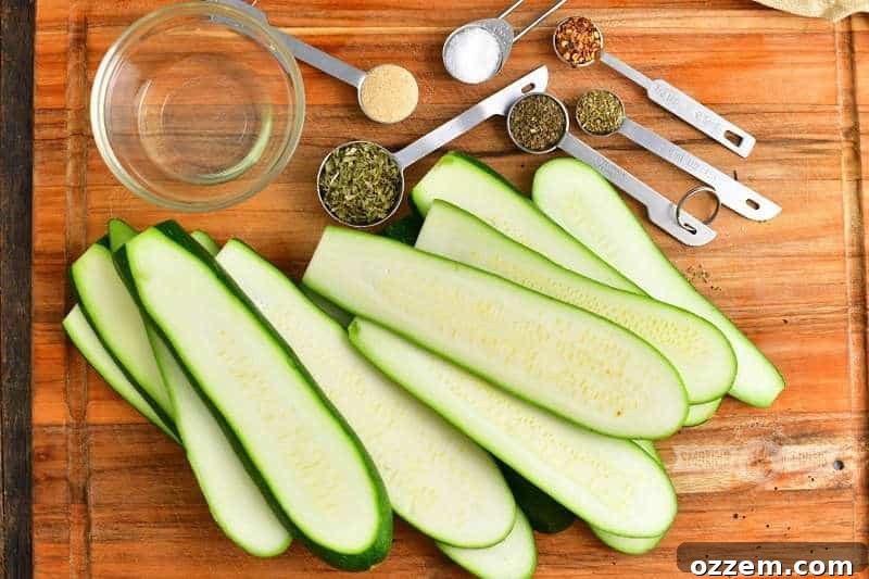 Sliced raw zucchini laid out, with a small bowl of olive oil and individual measuring spoons holding dried parsley, basil, oregano, salt, garlic powder, and crushed red pepper flakes on a rustic wooden cutting board.