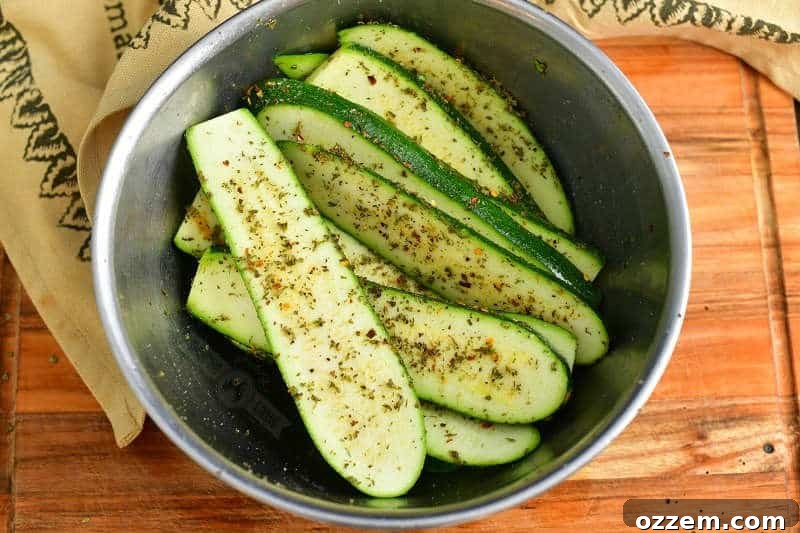 Long slices of zucchini, perfectly coated with olive oil and a savory seasoning blend, resting in a large mixing bowl on a wooden cutting board, with a brown linen towel draped nearby.