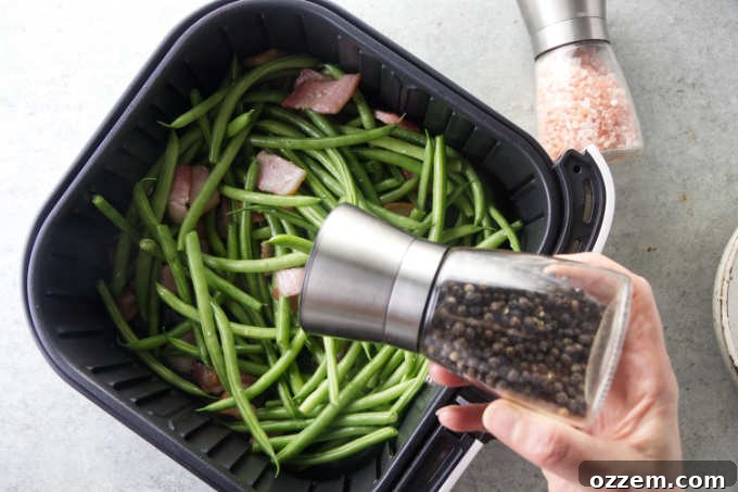 Adding black pepper to green beans and bacon already in the air fryer basket.