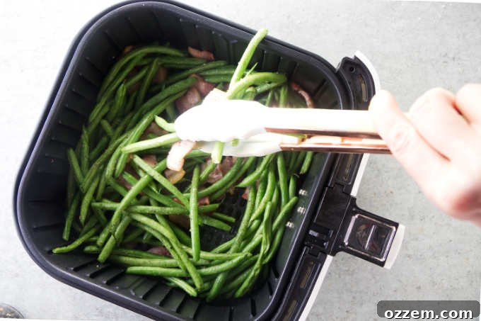 Stirring the green beans and bacon in an air fryer for even cooking.