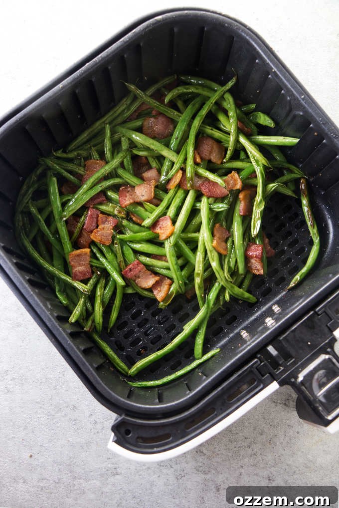 Crispy green beans and bacon cooking together in an air fryer basket, steam rising.
