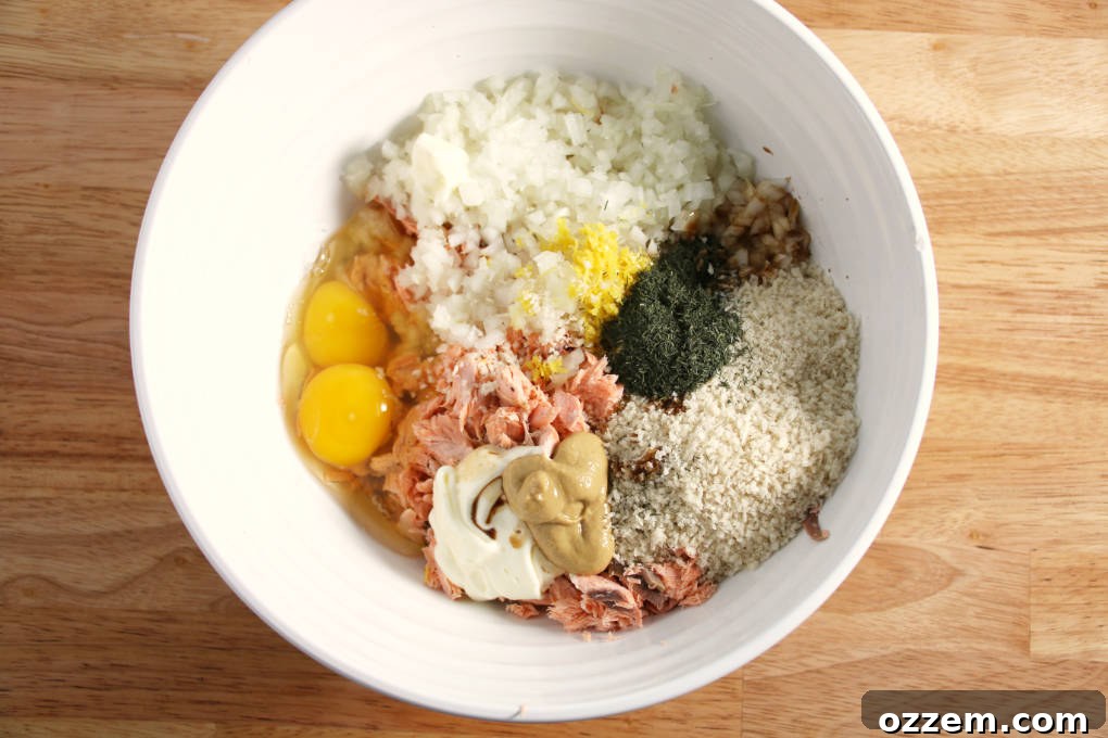 A stainless steel bowl filled with all the raw ingredients for air fryer salmon cakes, awaiting mixture. Features flaked salmon, diced onion, Panko breadcrumbs, eggs, lemon, and spices.