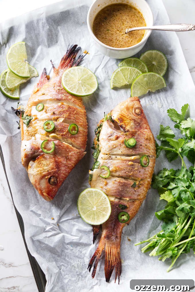 Perfectly grilled whole tilapia resting on a tray, garnished with fresh lime slices, herbs, and a vibrant bowl of chili-lime sauce.