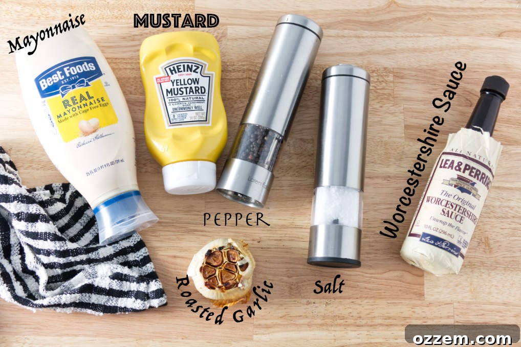 A beautiful flat lay of all the essential Donkey Sauce ingredients: a bowl of creamy mayonnaise, a roasted garlic bulb, a bottle of Worcestershire sauce, yellow mustard, and small dishes of kosher salt and freshly ground black pepper.